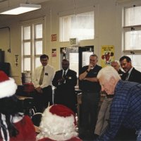 12-20-02 - Mission Education Center Christmas with Santa, Mission Education Center, San Francisco - Al Gentile, front right, hands Santa another present. Members looking on, L to R starting by the door, are Sheriar Irani, Dr. Mehendra Dave, Aaron Straus, Dick Johnson, Steve Currier, and Joe Farrah. Deborah Molof, Principal stands in the back left.