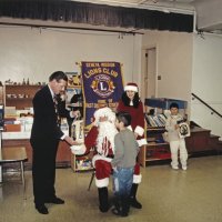 12-20-02 - Mission Education Center Christmas with Santa, Mission Education Center, San Francisco - Santa’s assistant look on while Steve Currier hands Santa the students gift bag while he waits anxiously waits. A student, happy with his gift, waits for his friend to receive his gift in the background.