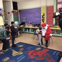 12-15-16 - Christmas with Santa with the help of Los Bomberos Firefighters, Mission Education Center, San Francisco - Santa listens intently as a students tells him what they’d like; a Los Bomberos Firefighter waits to hand Santa a gift for the student.