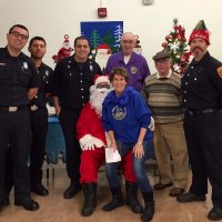 12/13/19 - Mission Education Center Christmas with Santa and Los Bomberos Firefighters - members of Los Bomberos Firefighters, Lion Aaron Straus (purple shirt) next to Lion Robert Lawhon, Geneva-Excelsior Lions member, Santa and Principal Deborah Molof. Lion Aaron was a member of the Mission Lions who founded this program more than 50 years ago.