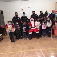 12-10-21 - Mission Education Center Christmas with Santa @ MEC, San Francisco - Students, with their gifts, pose with Santa and the Los Bomberos Firefighters. Their teacher is last on the right.